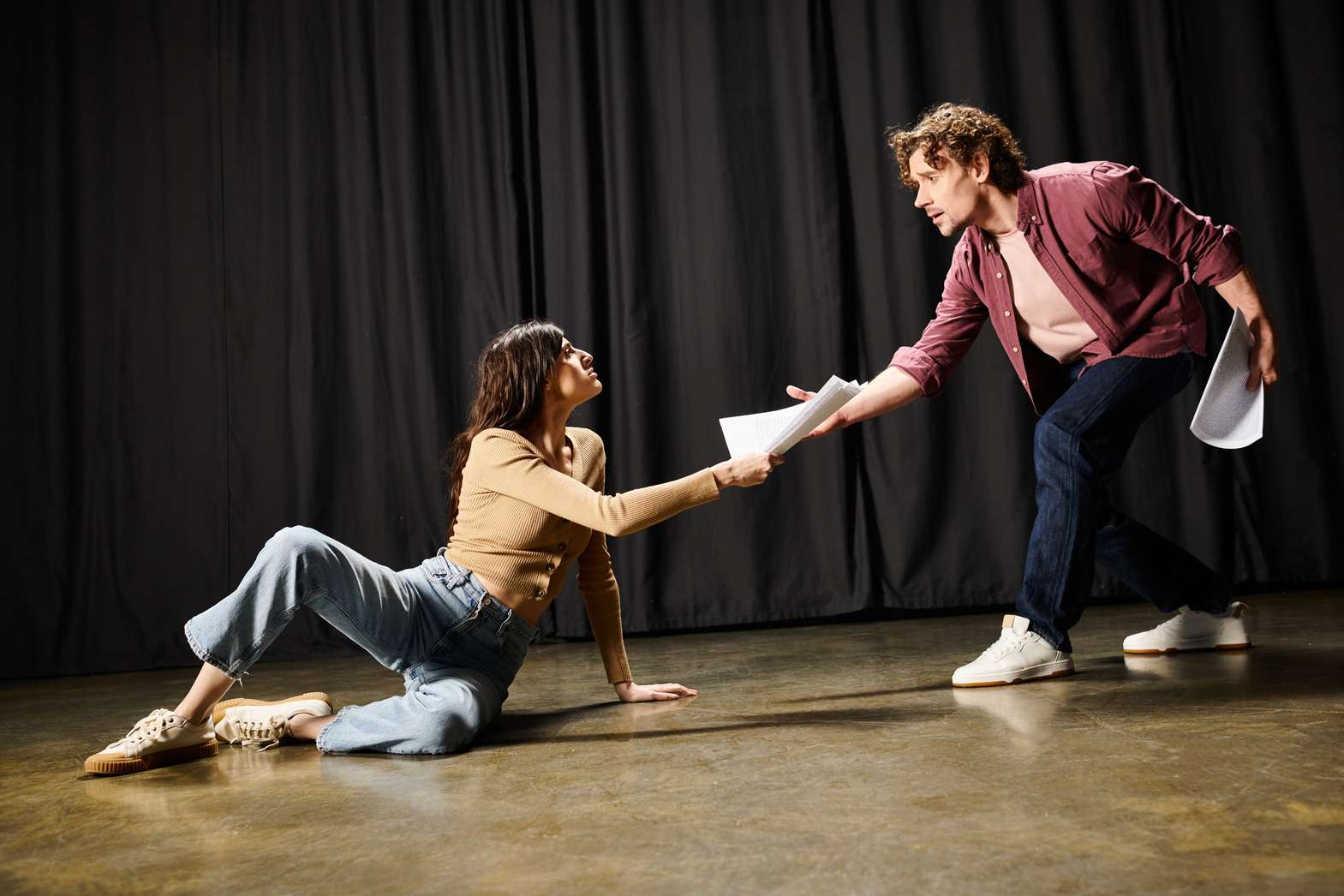 1. Un hombre y una mujer sentados en el suelo, él sostiene un papel mientras conversan. 2. Hombre y mujer en el suelo, uno de ellos sostiene un papel, compartiendo un momento juntos. 3. Una pareja sentada en el suelo, el hombre tiene un papel en la mano, ambos parecen estar en conversación.